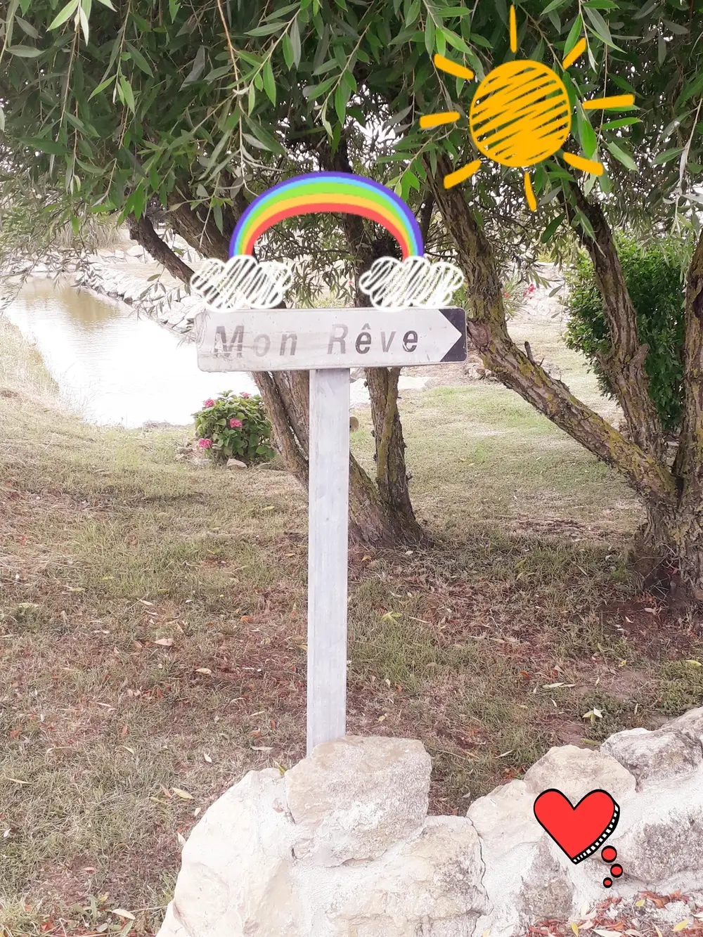 Au bord d'un chemin, un panneau de bois indique une direction : Mon Rêve. Un soleil et un arc-en-ciel sont dessinés par-dessus la photo.
