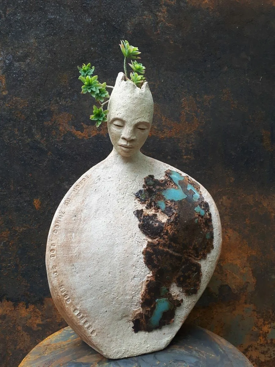 Une sculpture en céramique claire : un personnage aux yeux clos, le corps rond traversé d'éclats turquoise, des plantes qui poussent au sommet de sa tête.